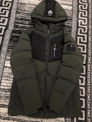 Grön pufferjacka från Sail Racing - Säljer en grön och svart pufferjacka från Sail Racing med huva och feta logodetaljer på bröstet och ärmen. Jackan har två bröstfickor med dragkedja, hel dragkedja framtill och quiltad design. Perfekt för kalla dagar och har en riktigt clean look. Jackan är vindtät. Pris kan diskuteras. Tveka inte på att skriva…