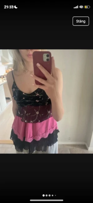 Blommig volang-linnetopp i svart och rosa - Söt linne-topp med tunna axelband och flera lager volanger. Överdelen är svart med broderade blommor i rosa och grönt, underdelen har rosa och svarta lager med spetsdetaljer. Luftigt material, perfekt för varma dagar och en trendig Y2K-vibe.