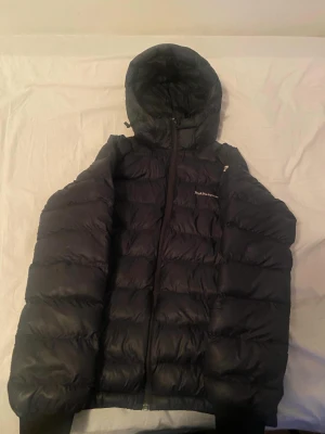 Svart Peak Performance dunjacka - Säljer en svart dunjacka från Peak Performance med huva och lång ärm. Jackan har en quiltad design och dragkedja framtill. Perfekt för kalla dagar och har en clean, sportig look som funkar till streetwear. Jackan är i storlek 170.