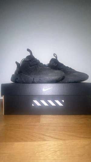 Nike React helsvarta sneakers - Säljer ett par helsvarta Nike React sneakers med snygg futuristisk design och chunky sula. Ovandelen är i mesh och syntet med diskreta reflexdetaljer och klassisk snörning. Perfekta för dig som gillar streetstyle och vill ha bekväma skor med cool look.