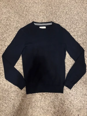 Mörkblå stickad tröja från Pier One - Snygg mörkblå stickad tröja från Pier One med rund hals och diskreta vita ränder på insidan av kragen. Tröjan har långärmade och ribbade muddar vid ärmslut och nederkant. Perfekt för dig som gillar stilrena och klassiska plagg.