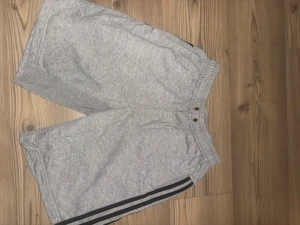Grå Adidas shorts med svarta ränder - Säljer ett par gråa Adidas shorts med klassiska svarta ränder längs sidorna. De har elastisk midja med snörning och är tillverkade i mjuk bomull, perfekta för chill dagar eller träning. Sportig och avslappnad stil.