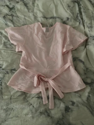 Ljusrosa omlottopp från Gina Tricot - Säljer en ljusrosa omlottopp från Gina Tricot med korta ärmar och v-ringad hals. Toppen har ett knytband i midjan som ger en snygg siluett och är tillverkad i mjuk bomull. Perfekt för dig som gillar feminina detaljer och enkel stil. Helt ny och aldrig använd