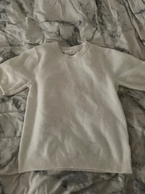 Vit stickad tröja från H&M - En enkel och clean vit stickad tröja från H&M med rund hals och korta ärmar. Tröjan har ribbade muddar vid hals och nederkant, vilket ger en snygg och stilren look. Perfekt för lager-på-lager under hösten eller våren.