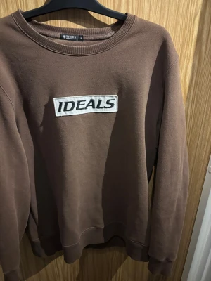 Brun sweatshirt Tiger of Sweden - Brun sweatshirt från Tiger of Sweden i storlek XL med broderad vit patch och svart text 'IDEALS' på bröstet. Tröjan har rund hals, ribbade muddar och är långärmad. Perfekt för dig som gillar stilrena och enkla plagg med en cool detalj.