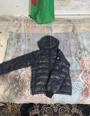 Svart dunjacka Peak Performance - Snygg svart dunjacka från Peak Performance med huva och dragkedja framtill. Jackan är quiltad och har ribbade muddar vid ärmsluten. Perfekt för kyliga dagar och har en stilren look som passar till det mesta. Priset kan diskuteras men de plick taxing