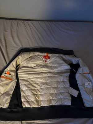 Svart quiltad jacka från Parajumpers - Säljer en svart quiltad jacka från Parajumpers med hög krage, dragkedja framtill och flera fickor. Jackan har ribbade muddar och detaljer i orange. Materialet är polyester och fleece, perfekt för kyligare dagar.