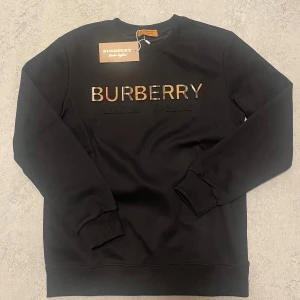 Svart Burberry sweatshirt med logga - Svart sweatshirt från Burberry med klassisk rund hals och ribbade muddar. Framsidan har Burberrys logga i relief med rutiga detaljer i bokstäverna. Tröjan är tillverkad i mjuk bomull och har en clean, stilren look som passar perfekt till jeans. Byten funkar.