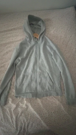 Grå hoodie med dragkedja och huva - Säljer en Jack n Jones grå hoodie med dragkedja framtill och två fickor. Tröjan har huva och långa ärmar, perfekt för chill dagar. Materialet känns mjukt och skönt, troligen bomull eller polyester. Passformen är normal och färgen är neutral grå.