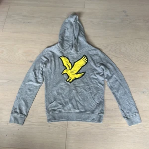 Grå hoodie med gul fågel från Lyle & Scott - Säljer en grå hoodie från Lyle & Scott med en stor gul fågel på bröstet. Tröjan har huva, magficka och långa ärmar. Perfekt för dig som gillar sportig och avslappnad stil. Materialet är mjukt och skönt bomullsmix. Aldrig använd