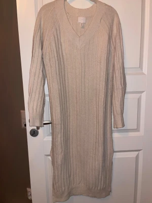 Beige ribbstickad klänning H&M - Supermysig beige ribbstickad klänning från H&M med v-ringning och långa ärmar. Perfekt för lager-på-lager och chill dagar. Mjuk och skön, med en lite längre modell som ger en avslappnad vibe.
