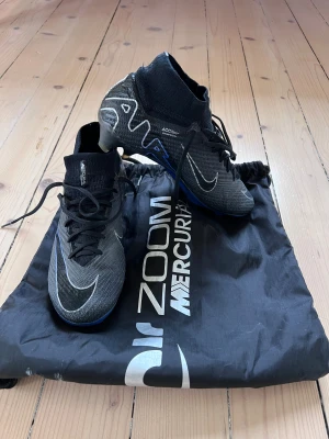 Nike Mercurial Zoom fotbollsskor svart - Säljer ett par Nike Mercurial Zoom Elite fotbollsskor i svart med blå detaljer och vita loggor. Skorna har strumpliknande skaft, snörning och en lätt, syntetisk ovandel för bästa känsla på planen. Perfekta för dig som vill ha snabbhet och kontroll. Nypris 2700:-