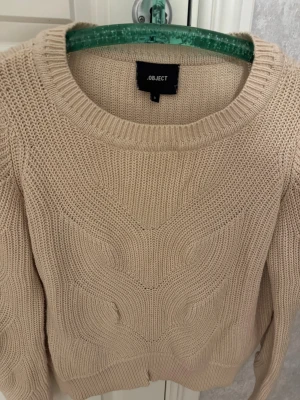 Beige stickad tröja från Object - Snygg beige stickad tröja från Object med rund hals och coolt strukturerat mönster framtill. Tröjan har långa ärmar och ribbade muddar vid hals, ärmslut och nederkant. Perfekt för dig som gillar neutrala färger och sköna material.