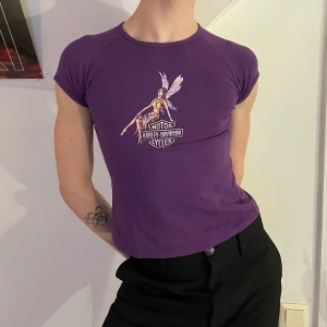 Lila vintage y2k graphic babytee älva - Lila vintage Harley Davidson babytee med ett tryck av en älva som är smått glittrig 🧚‍♀️ Eftersom den är stretchig är den så skön och passar perfekt till ett par stora jeans eller cargobyxor!