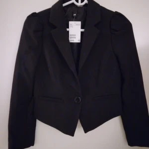 HELT NY & OANVÄND snygg svart kort kavaj / jacka. Strlk 34 / XS.  - Helt ny och oanvänd svart snygg kort kavaj med snygga ärmar med lite rynk och puff på. Stängs med knapp. Tags sitter kvar sedan inköpet. Material: Polyester. Satinfoder. Storlek: 34 / XS. Helt felfri. FRAKT TILLKOMMER.