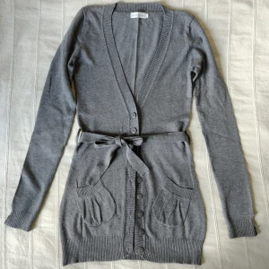 Gråmelerad längre knäpptröja/kofta med knytband, storlek 8 - Gråmelerad längre knäpptröja med avtagbart knytband i midjan. Tröjan är mjuk och elastisk. Sparsamt använd. 49% viskos 27% bomull och 24% nylon. Storlek 8, passar XS/S. Något smånopprig/luden på vissa ställen. 