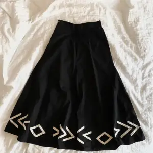  En svart midjehög kjol med vita detaljer längst ner.  Dragkedja och spänne på sidan.   Mycket bra skick.