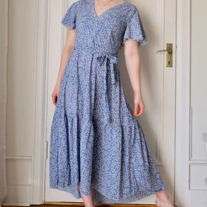 Blå långklänning i storlek 36 - Blå långklänning med vita blommor på i storlek 36. Modellen på bilden är 159 cm. Härlig sommarklänning med band att knyta runt midjan. Klänningen är i nyskick- aldrig använd. 
