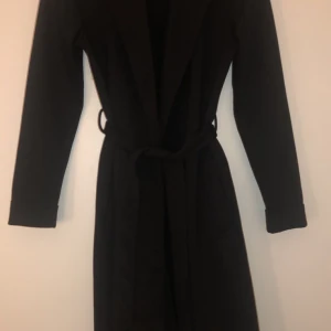 Svart kappa - Det är en svart kappa med två fickor framme, det finns ett ”rep” i midjan. 