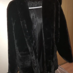 Fuskpäls - En fuskpäls som är oanvänd. Man knäppar jackan i dolda hättor.  Med luva och 2 stora fickor