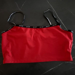 Röd topp med svarta axelband från Levis, strl. M - Röd magtröja/topp i glatt material från Levis, strl M. Väl använd men i bra skick. Köparen står för frakt🌻