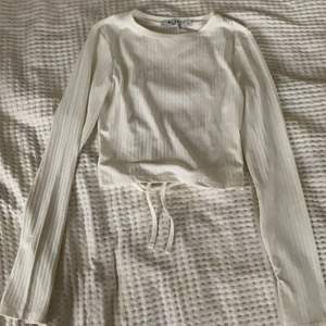 En ribbstickad vit lång tröja med långa ärmar så man kan ha de över handlederna, öppen i ryggen med knytning. I strlk xs