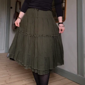 mörkgrön volangkjol - En mörkgrön/kakigrön volangkjol från Prêt á Porter. Fint skick. Kjolen har en tunn underkjol i samma färg och ett knytsnöre i midjan. Strl 36 men passar även 34 då man kan knyta åt hur man vill. Jag som bär kjolen är 160cm och bär S/36. Längd: 65cm. Midjemått: 68-86cm.  