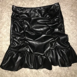 Festlig Svart volang kjol  - Kjol i pu läder . Storlek 38 . Helt ny, inte kommit till användning. Dragkedja på sidan. 