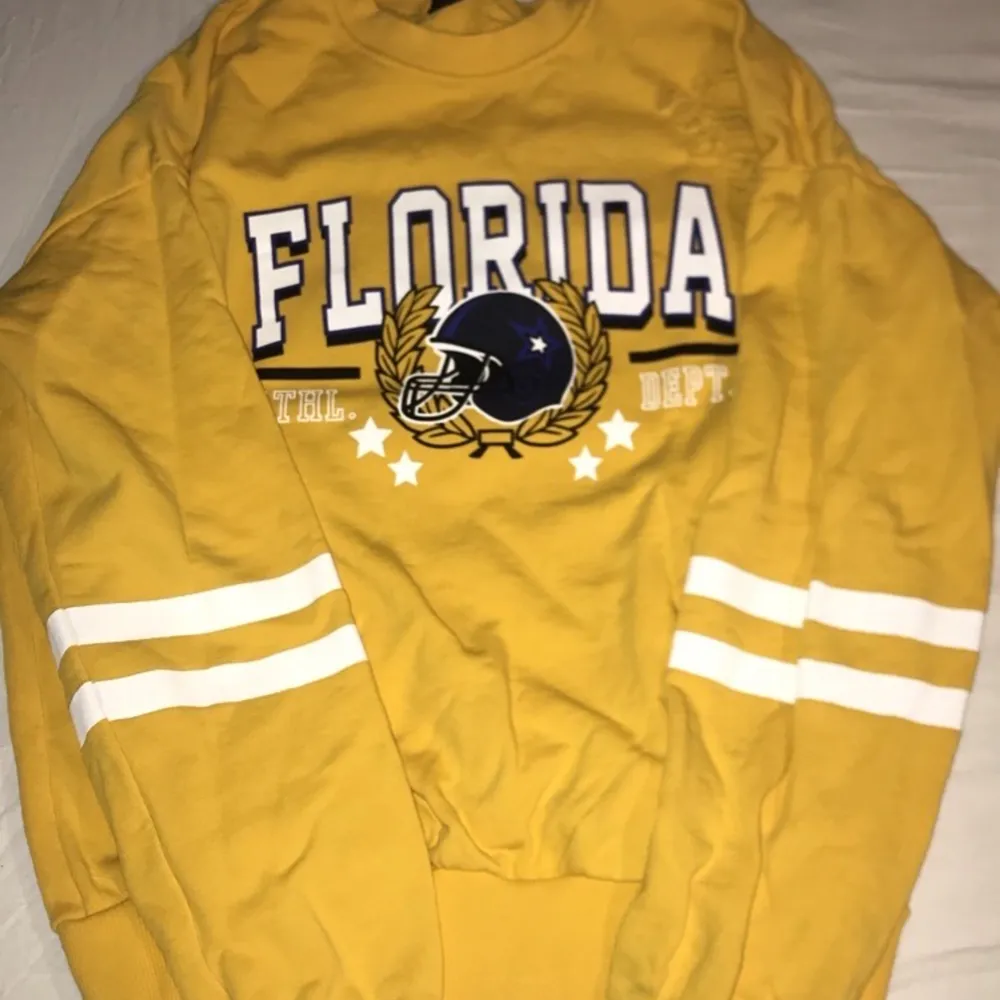 En gul långärmad tröja med trycket ”Florida” tvärs över bröstet. Det är ”slitningar” över hela tröjan samt två vita sträck på båda ärmarna. Storleken är 36/S. . Tröjor & Koftor.