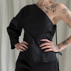 Assymetrisk topp - Enarmad topp i svart glansigt material från Weekday. Figursydd, knäpps med tryckknappar och snöre på en sida. Sååå snygg!! Ärmen är lite ballongig utan att vara stor. Passar S-M