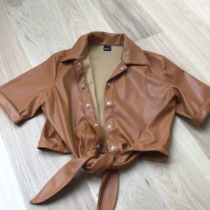 Gina Tricot top läder - Jätte bra skick inte skador eller något liknande. Hasselnötsbrun man kan knytta den på många olika sätt och knäppa upp hur mycket man vill. Alla knappar finns. Från Gina Tricot 