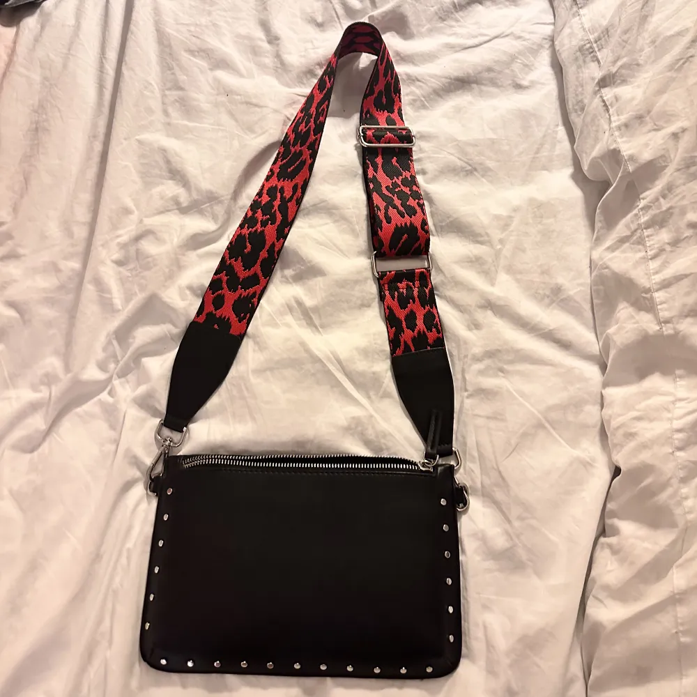 Liten svart väska med silvriga detaljer och ett rött leopardmönstrat band som är justerbart❤️. Väskor.