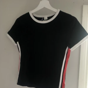 Svart t-shirt med röd/vita ränder - Svart t-shirt med röd/vit rand på sidan av tröjan. Från h&m. Använd men fåtal gånger. 