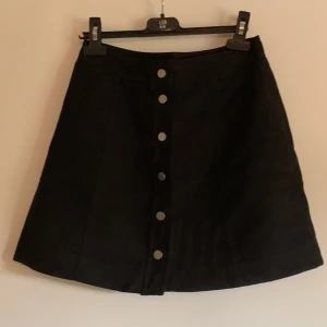 Kjol i suede-imitation - Svart kjol i ett tyg liknande suede med knappar längs framsidan. Alla knappar går att knäppa upp. Använd en del men i väldigt gott skick! Storlek 34. 