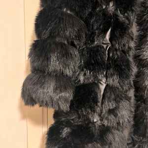 Svart fuskpäls kappa - En elegant och fyllig jacka av fuskpäls med en stor och fin luva. Minns ej nypriset eller butik. Passar xs. Använd några gånger! Fortfarande som ny. 