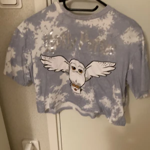 Harry Potter merch - Harry Potter t-shirt från hm. Det står Harry Potter med silverfärgad och hans Uggla Hedwig på tröjan. Den säljs inte längre. Den är bred och kort och stor i storleken men ingen magtröja direkt. Den är ner till armbågarna. Den är blå och vit.