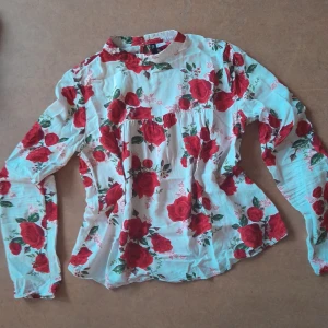 Blus med röda rosor - En vit blus med röda rosor. Långärmad och knäppning i nacken. Storlek 36 från H&M Divided, och i mycket fint skick. Skönt och svalt tyg.