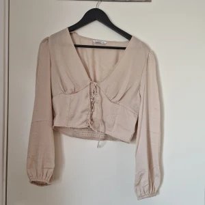 Blus i satinmaterial - Beige satinblus med snörning. Storlek L men liten i storleken. Passar storlek S och M. Endast använd en gång. Köpt på Asos. Nypris 289 kr.