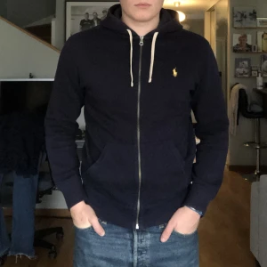 Ralph lauren tröja - Mörkblå ralph lauren hoodie med dragkedja. Den är i bra skick förutom den vänstra armbågen som syns på bild 2. Nypris: 1800kr  Mitt pris: 500kr.  Modellen på bilden är 184cm och väger 90kg.