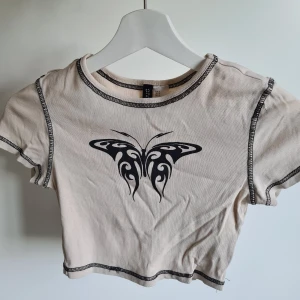 Crop top - En beige magröja med en svart fjäril på och svarta sömmar