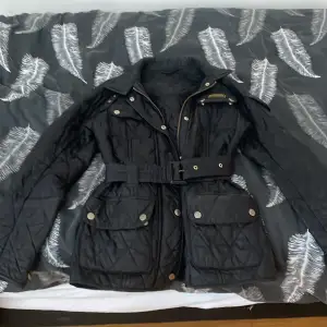 En svart äkta Barbour jacka som passar xs,s och m. Med ett bälte och tragkedjor vid händerna samt fyra fickor   