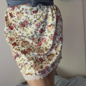 Blommig kjol i mjukt material som liknar silke - Blommig kjol i mjukt material som liknar silke. Den har öven en underkjol med spets i kanten. Kjolen sluta strax över knäna.Aldrig använd, endast provad