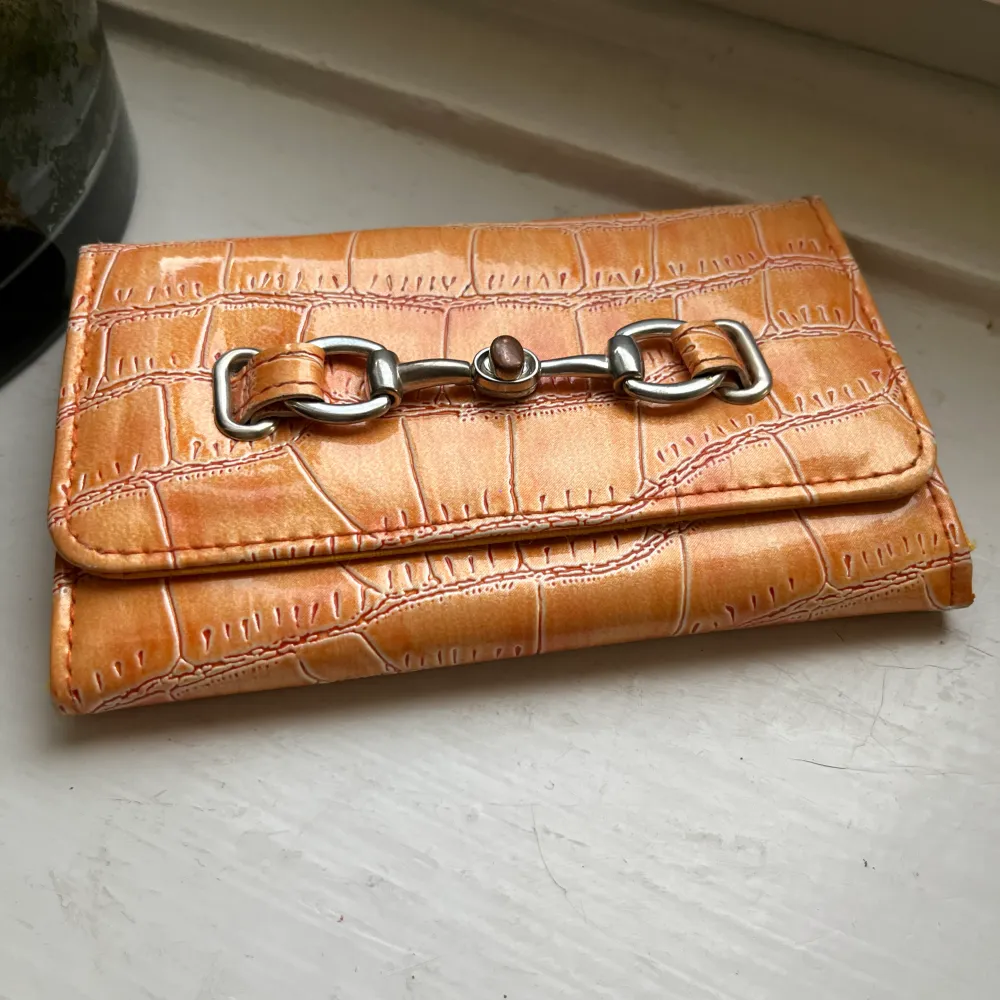 En orange plånbok som jag inte var vart den kommer ifrån. Den har en silver ditalj där framm och har mönster.  Kommer sälja pga inte använder lika mycket som förut! . Väskor.