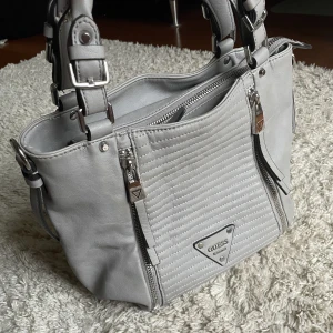 Guess väska - Guess väska. ❤️ Går att ha som både handväska eller axelväska. Rymlig och har fyra fickor på insidan och även en på utsidan, har snygga silvriga detaljer med lyxigt mönster på insidan. 🫶 Håll koll på min profil, säljer för det mesta vintage kläder. 😍