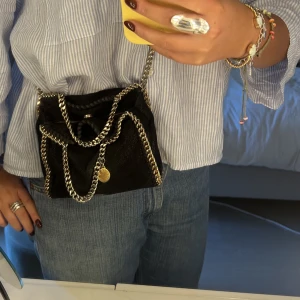 stella mccartney väska - Tänkte sälja denna väska från Stella mccartney. En svart med guldig kedja. Den är i fint skick förutom att lite färg lossnat från den guldiga cirkeln. Detta är den minsta modellen.