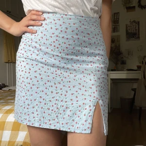 Kort kjol med slits - Ljusblå kort kjol med röda blommor som stängs med dragkedja i sidan. Jättefin men kommer aldrig till användning. En del av fodret är löst men det går att fixa med säkerhetsnål, eller om man vill sy. 