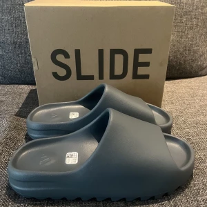 YEEZY SLIDE SLATE MARINE - Yeezy Slide Slate Marine. Helt oanvända, storlek 44 1/2. Små i storleken, passar till dem som har 42 - 43. Köpta från Adidas Confirmed appen så kvitto mm finns.