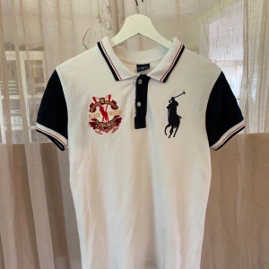 Vit och marinblå pikétröja från Polo Sport Ralph Lauren - En stilren vit och marinblå pikétröja från Polo Sport Ralph Lauren. Tröjan har korta ärmar, en krage med knappar och broderade detaljer på bröstet. Den är tillverkad i ett bekvämt material och passar perfekt för både vardag och sportiga aktiviteter.