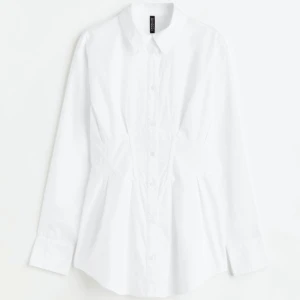 Poplinskjorta med intagen midja - En något längre skjorta i bomullspoplin med spetsig krage och knäppning fram. Skjortan har avslappnad passform, lätt nedhasad axel och lång ärm med manschett som har knäppning. Intagen midja med lagda veck och fällsöm fram. Aldrig använts,prislapp på