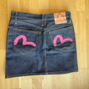 Jeanskjol från evisu - Helt ny jeanskjol från evisu med rosa detaljer där bak! Har inte använts en ända gång. Kjolen är 100% av bomull. Den är ganska liten i storleken så jag skulle säga att den passar S/XS.  Längd: 40 cm Midjemått tvärs över: 37 cm